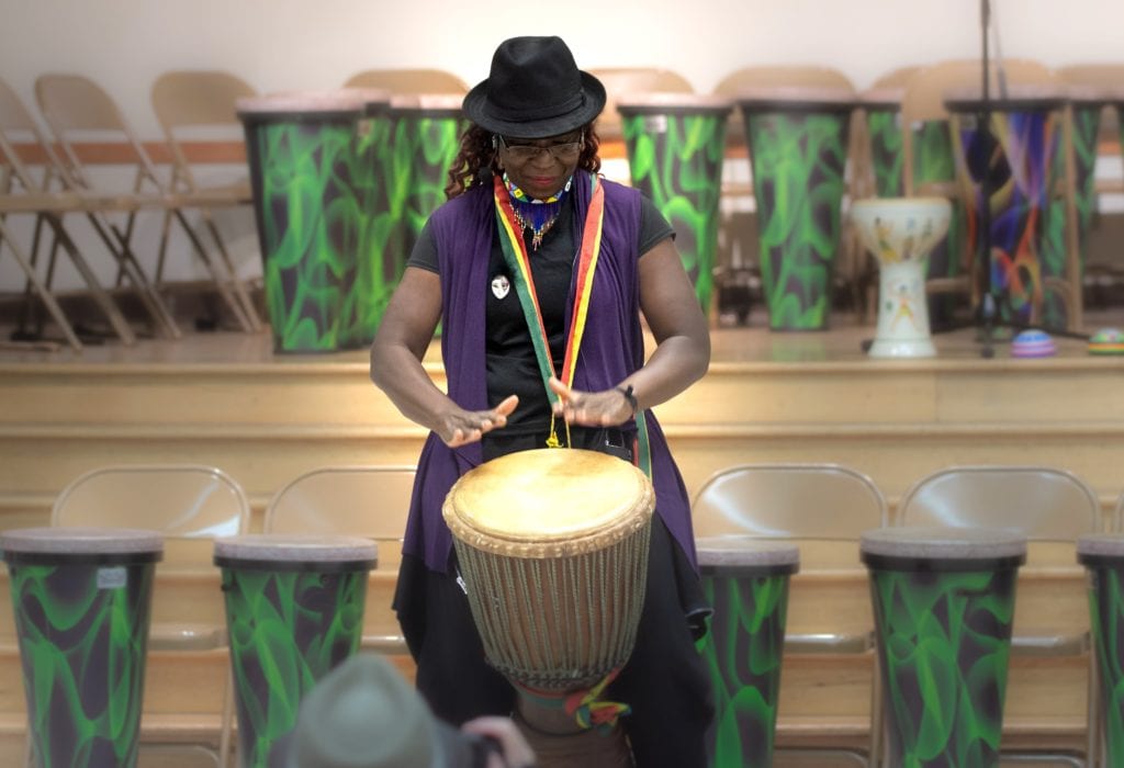 Black woman with a rainbow lanyard plays a drum in an auditorium.