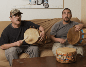 two men sitting on a couch playing the drums