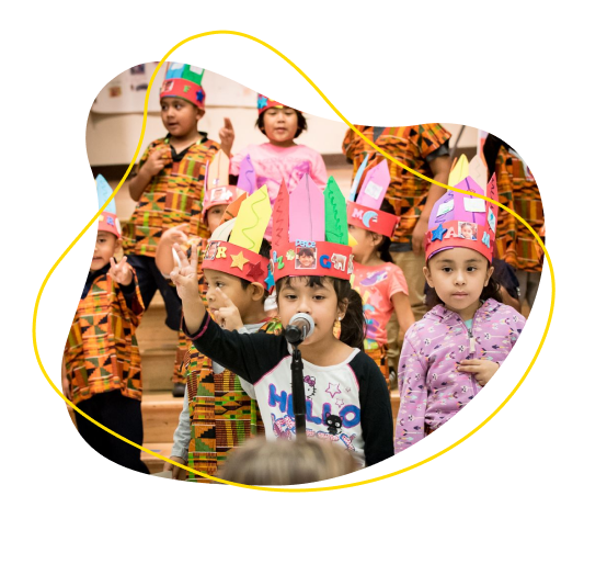 Children wearing construction paper Native American headdresses and standing before a microphone.