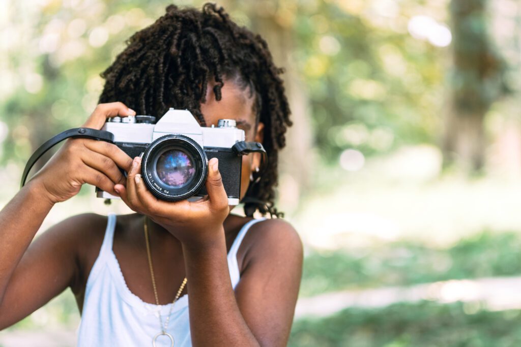 Young black girl points a DSLR camera outside on a sunny day in a photography class.