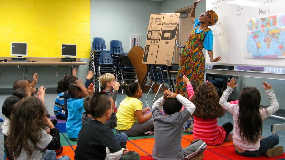 A teacher standing in front of a dry erase board as childern look on while sitting on a carpeted area.