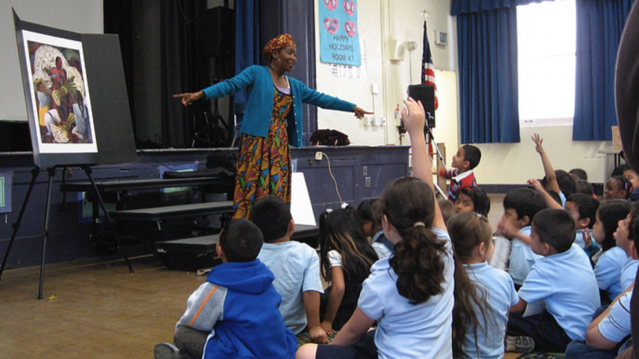 A black teacher acting in front of a seated group of children in a theatre class.