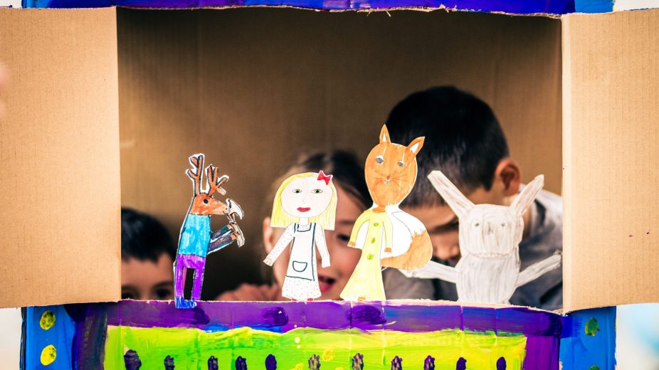 Close up of children playing with paper puppets in a painted cardboard box.
