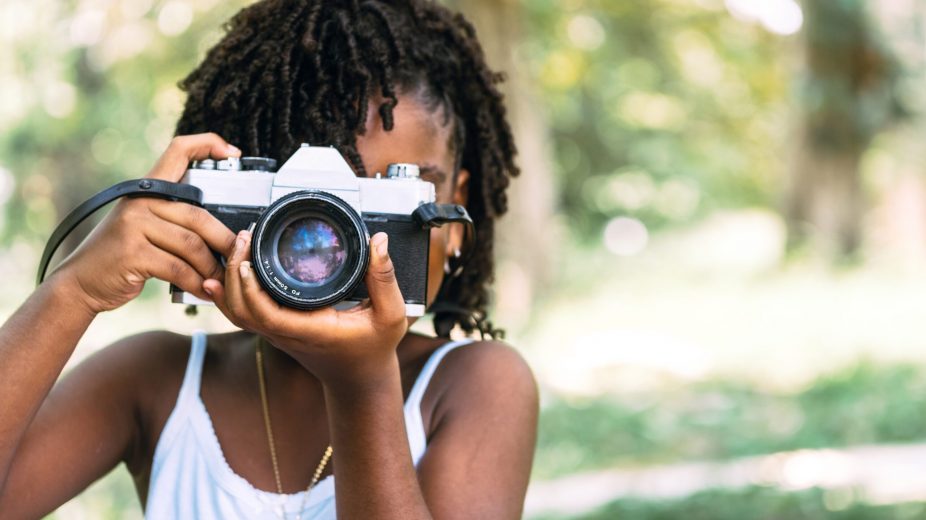 Young black girl points a DSLR camera outside on a sunny day in a photography class.